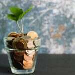 Coins in a clear glass container with a small green plant growing out of the top, symbolizing gradual financial growth and mindful money habits.