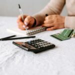 Person writing in a notebook beside a calculator, cash, and credit cards