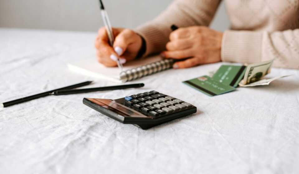 Person writing in a notebook beside a calculator, cash, and credit cards