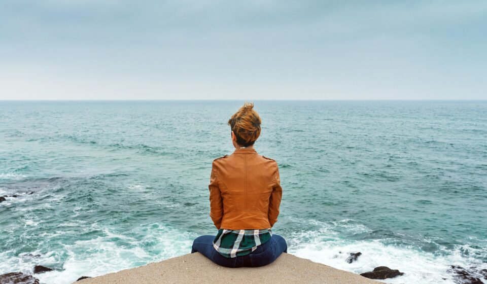 Woman sitting on a rock by the sea, looking out over the water on a quiet day.