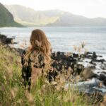 A person sitting on a rock by calm water, looking out toward the horizon in a quiet natural setting.
