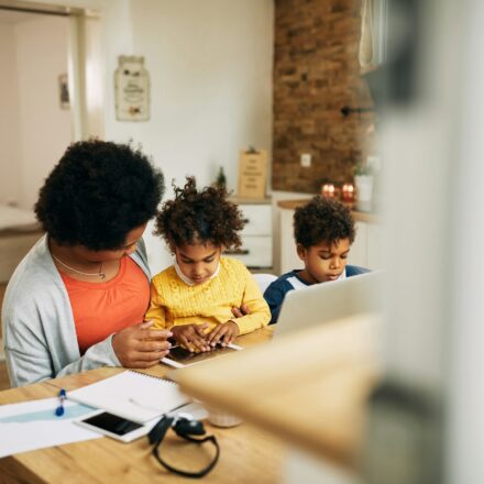 Mother working at a laptop at home while talking with her daughter as a young boy sits nearby