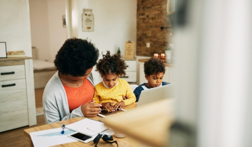 Mother working at a laptop at home while talking with her daughter as a young boy sits nearby
