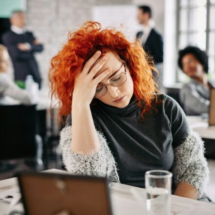Woman sitting at a desk with her head in her hands while coworkers move in the background.