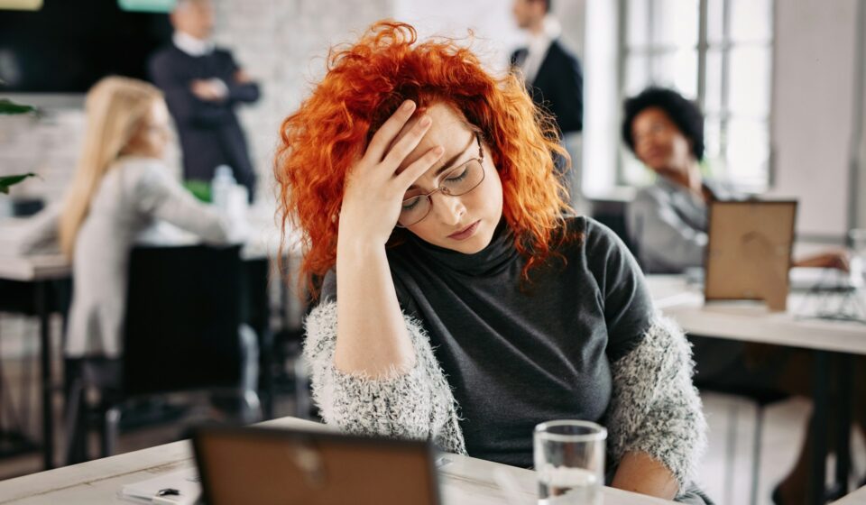 Woman sitting at a desk with her head in her hands while coworkers move in the background.