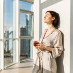 Woman standing near a sunlit window holding a mug in a calm home environment