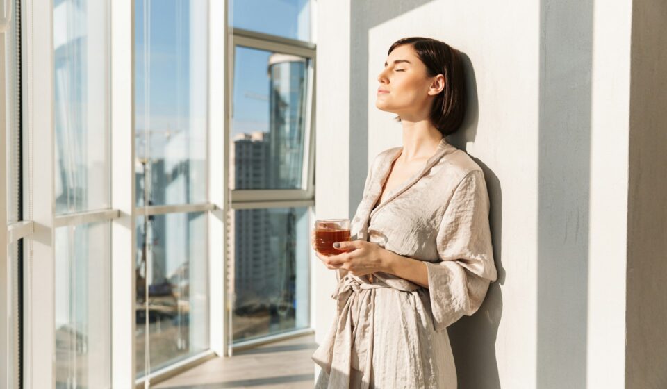 Woman standing near a sunlit window holding a mug in a calm home environment