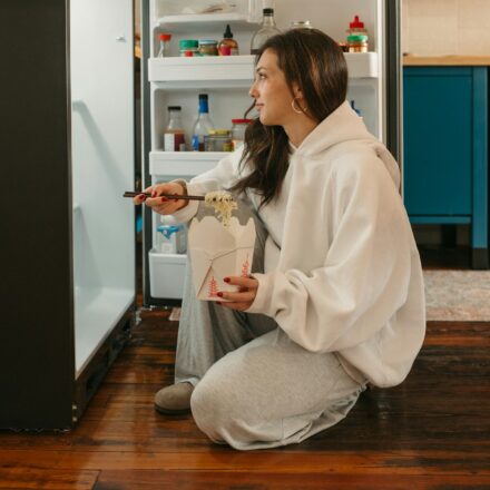 Woman standing at open refrigerator holding leftovers while deciding what to eat at home