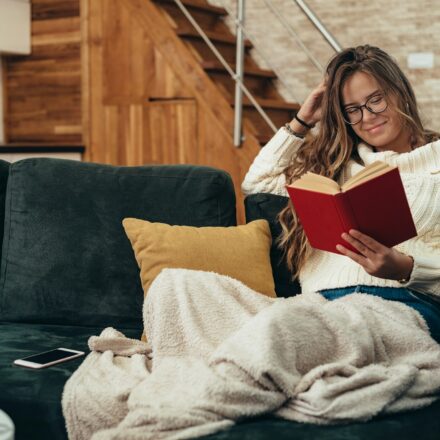 Woman relaxing on a couch reading a book with a smartphone nearby