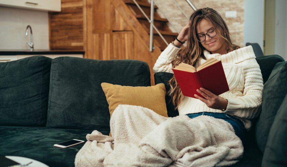 Woman relaxing on a couch reading a book with a smartphone nearby