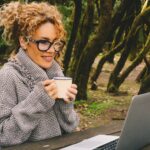 Woman smiling while working on a laptop outdoors among trees, representing focus, mental clarity, and a sense of ease during the day.