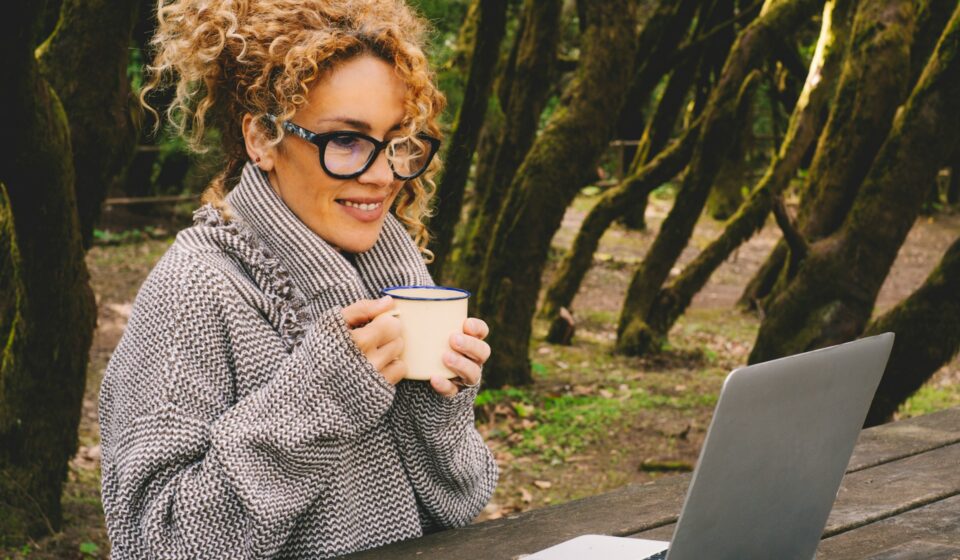 Woman smiling while working on a laptop outdoors among trees, representing focus, mental clarity, and a sense of ease during the day.