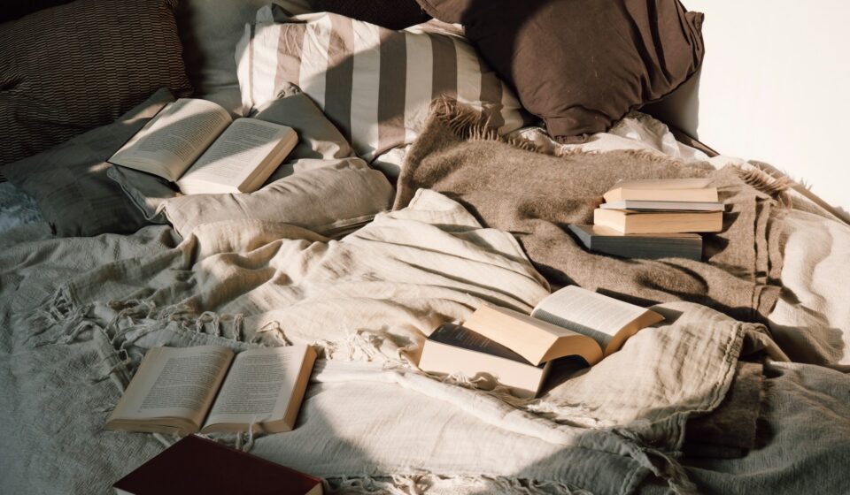 Cozy bed covered with blankets and cushions, with several open books scattered across the quilted bedspread.