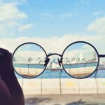 Black-framed eyeglasses held up against a city skyline with the view appearing clear through the lenses and blurred outside them.