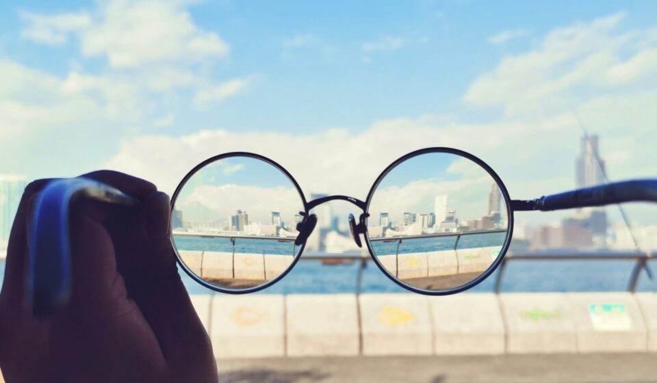 Black-framed eyeglasses held up against a city skyline with the view appearing clear through the lenses and blurred outside them.