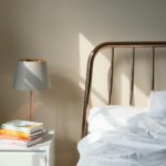 White nightstand with a lamp, a small succulent, and a stack of colorful books beside a copper bed frame and white bedding.