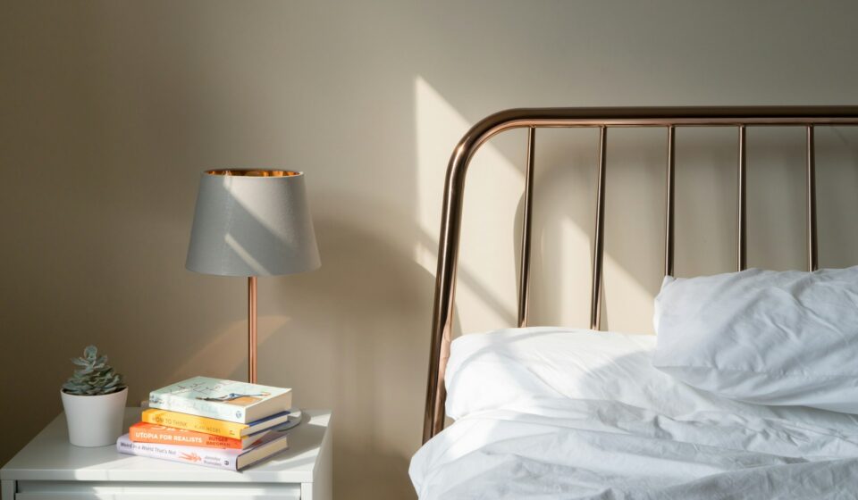 White nightstand with a lamp, a small succulent, and a stack of colorful books beside a copper bed frame and white bedding.