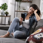 Woman sitting on a couch looking at her phone with a tired expression during a quiet moment at home