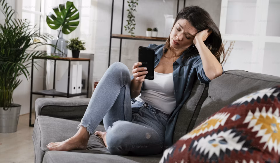 Woman sitting on a couch looking at her phone with a tired expression during a quiet moment at home